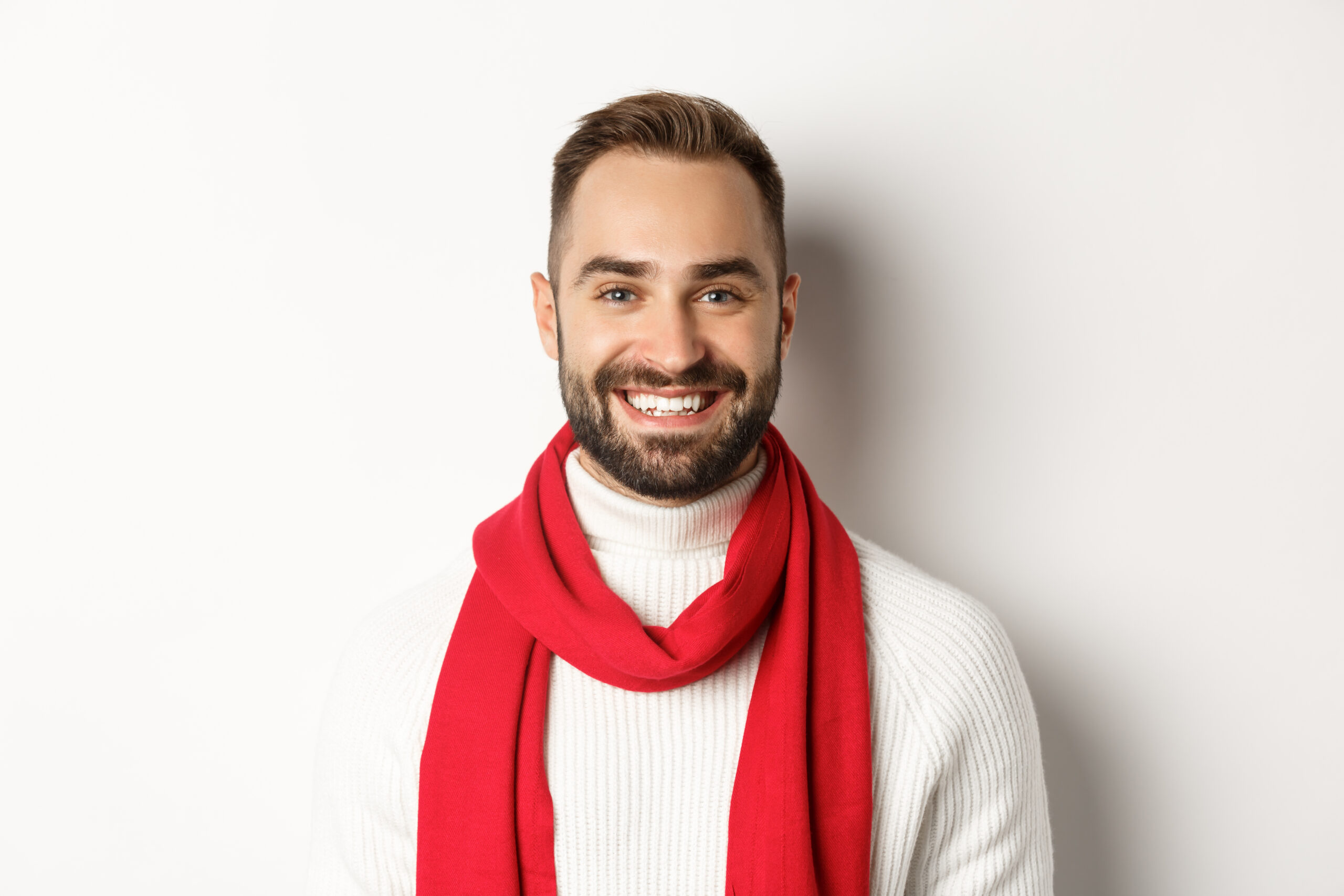 Christmas holidays. Handsome bearded man in red scarf and sweater smiling, white background.